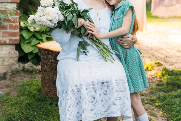 communication between grandmother and granddaughter. grandmother, an elderly woman in white, vintage clothes sitting and hugging her granddaughter, a girl in a green dress, and holding in her other hand a bouquet of flowers, white peonies, in the gar - Zdjęcie, obraz