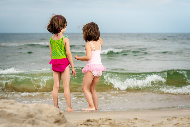 two little girls sisters are standing and playing on the sand at the beach on a summer day - Photo, Image