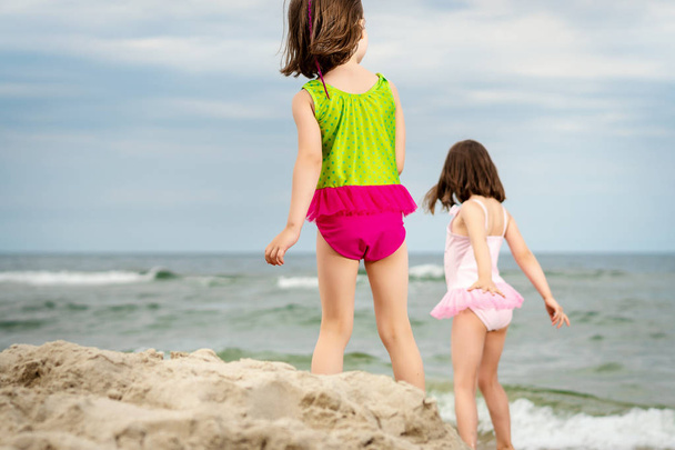 two little girls sisters are standing and playing on the sand at the beach on a summer day - Photo, Image