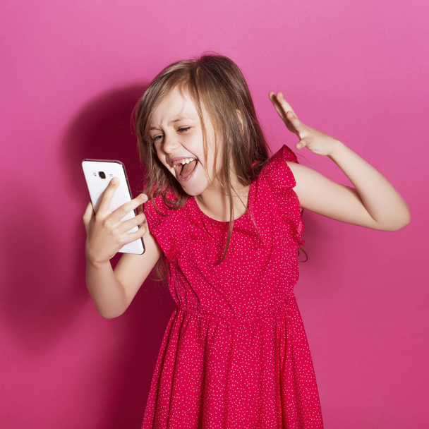 Little 8 years old girl make some emotional gesture with her hands on a pink neutral background. She has long brunette hair and wear red summer dress. Funny expression on her face - Photo, Image