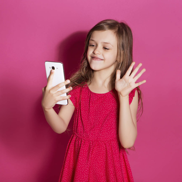 Little 8 years old girl make some emotional gesture with her hands on a pink neutral background. She has long brunette hair and wear red summer dress. Funny expression on her face - Fotoğraf, Görsel