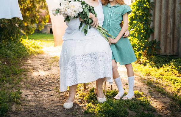 visit grandmother. grandmother, an elderly woman in white, vintage clothes sitting and hugging her granddaughter, a girl in a green dress, and holding in her other hand a bouquet of flowers, white peonies, in the garden. meeting and congratulations,  - Foto, Imagem