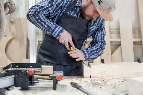 Un hombre moreno con barba y gafas verdes protectoras tratando un producto de madera con un cincel en el taller
 - Foto, Imagen