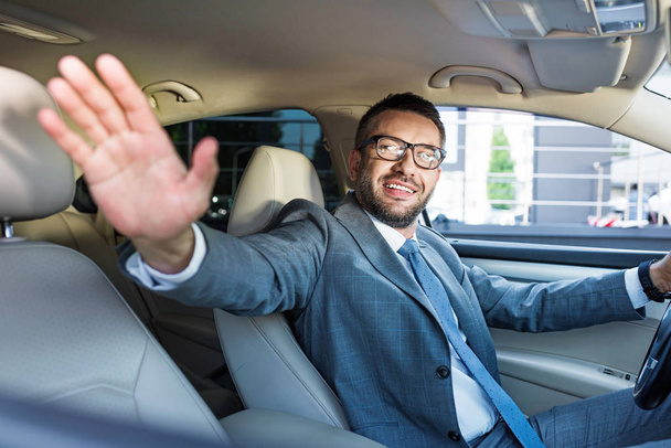 smiling businessman in suit and eyeglasses greeting someone while driving car - Zdjęcie, obraz