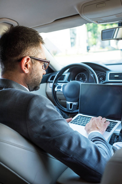 side view of businessman in eyeglasses using laptop with blank screen in car - Foto, afbeelding