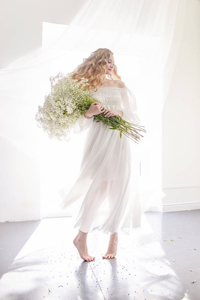 Curly blonde romantic look, beautiful eyes. White wildflowers in hands. Girl white light dress and curly hair, portrait of woman with flowers at home near the window, purity and innocence - 写真・画像