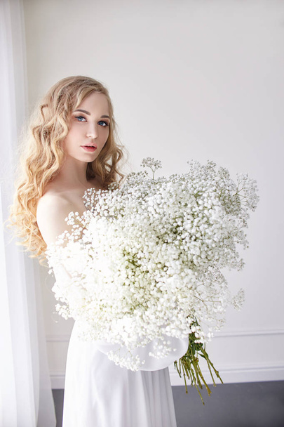 Curly blonde romantic look, beautiful eyes. White wildflowers in hands. Girl white light dress and curly hair, portrait of woman with flowers at home near the window, purity and innocence - Photo, Image