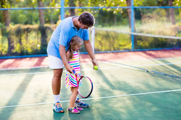 Father and daughter playing tennis on outdoor court. Family with tennis racket and ball at net in sport club. Coach teaching young kid. Child learning to play. Training for kids. Children exercise. - 写真・画像