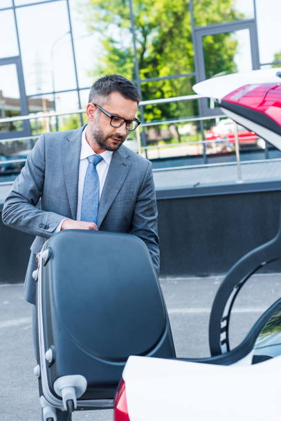 portrait of businessman in eyeglasses putting luggage into car on parking - Foto, Bild