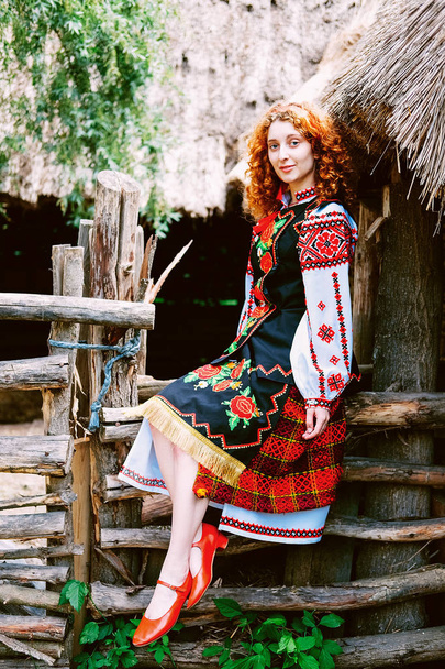junge slawische Frau in traditioneller bestickter Tracht und roten Schuhen auf der Veranda sitzend - Foto, Bild