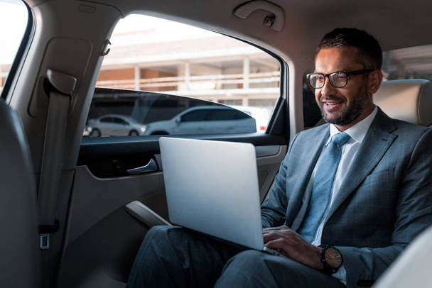 uśmiechający się biznesmen w okularach za pomocą laptopa na tylnym siedzeniu w samochodzie - Zdjęcie, obraz