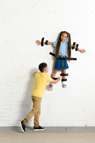 hermano pegando hermana a la pared con cinta negra en casa
 - Foto, imagen