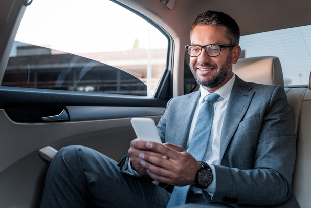uśmiechający się biznesmen korzystanie ze smartfona na tylnym siedzeniu w samochodzie - Zdjęcie, obraz