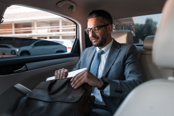 businessman in suit putting papers into bag while sitting on backseat in car - Fotografie, Obrázek