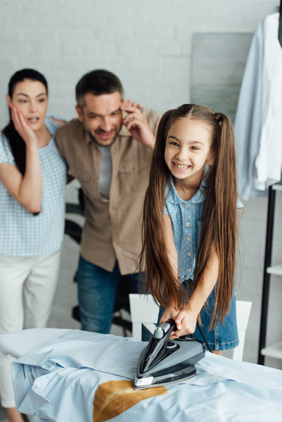 smiling daughter holding iron above burned shirt at home, parenthood concept - Photo, Image