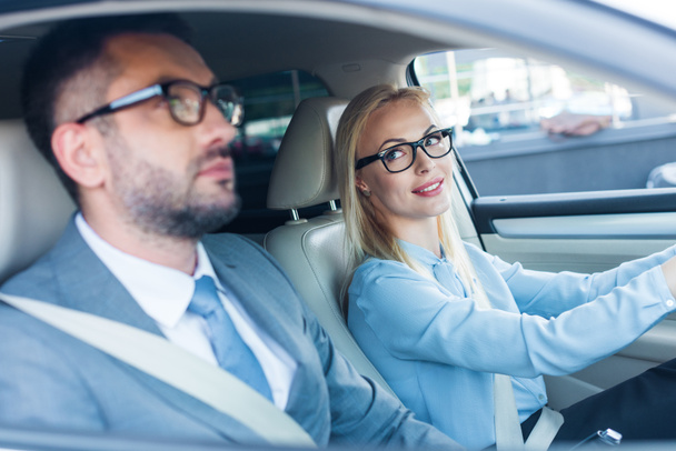 Zijaanzicht van blonde zakenvrouw in brillen rijden auto met collega in de buurt van door - Foto, afbeelding