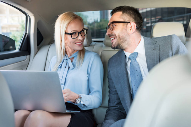 portrait of smiling business people with laptop on back seats in car - Фото, зображення