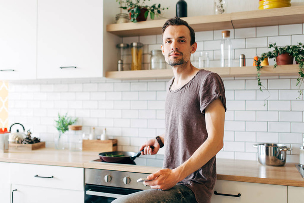 Portrait of brunet man with frying pan in his hands talking on phone in kitchen - Photo, Image