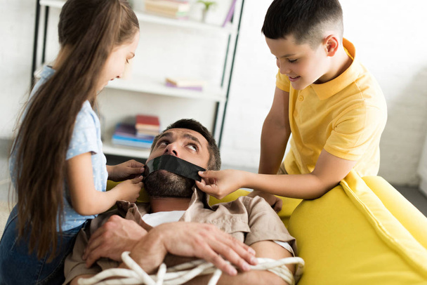 hijos desobedientes cerrando la boca del padre con cinta negra en casa, concepto de paternidad
 - Foto, imagen