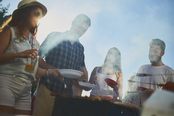 Groep mensen die staan rond grill, chatten, drinken en eten.  - Foto, afbeelding