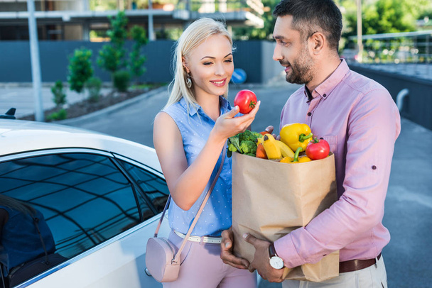 portrait of smiling married couple with paper bag full of healthy food on parking near car - Foto, immagini