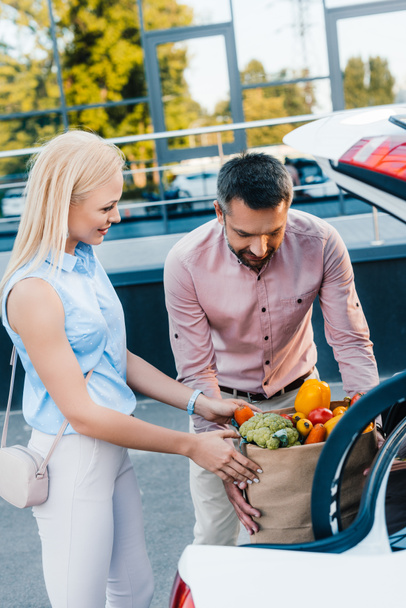 portrait of married couple putting paper bag full of healthy food into car on parking - Photo, Image