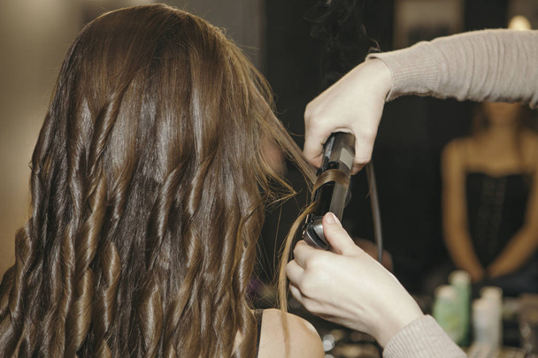 Maquillaje artista preparar a la mujer a su sesión de fotos. Se riza el pelo. Modelo sentada con su espalda a una cámara. Morena, pelo moreno
 - Foto, imagen
