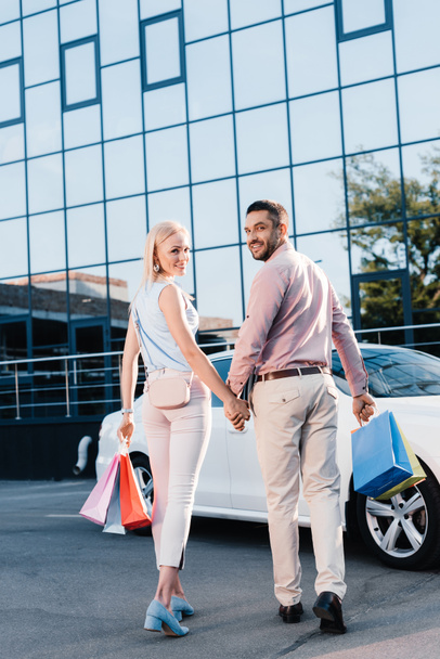 vista trasera de la pareja casada con bolsas de compras cogidas de la mano y caminando al coche en la calle
 - Foto, Imagen