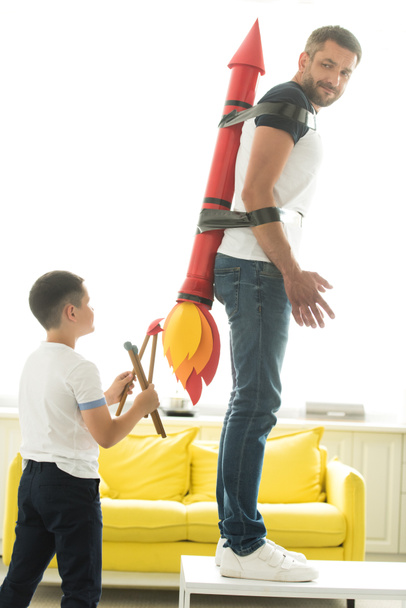Непослушный сын играет со связанным отцом с ракетной игрушкой дома
 - Фото, изображение