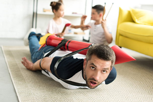 tied father with rocket toy lying on floor and children playing in living room - Photo, image