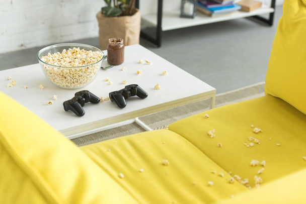 glass bowl with popcorn and joysticks on table, yellow sofa in living room - Фото, изображение