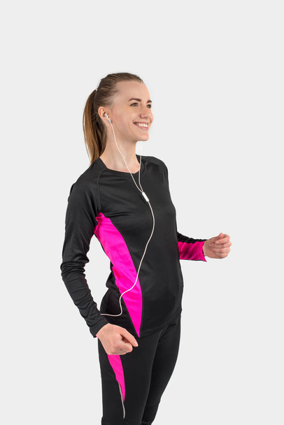 Усміхнена молода дівчина в чорному спортивному костюмі з рожевими смужками і з навушниками робить біг
 - Фото, зображення