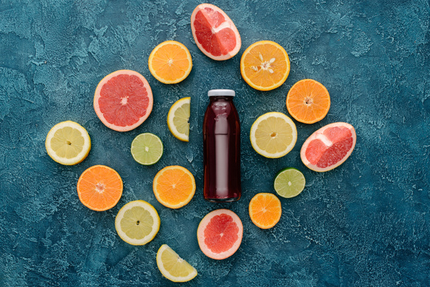 top view of bottle of red juice surrounded with citrus fruits slices on blue concrete surface - Photo, Image