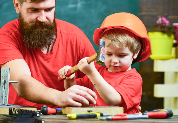 Little assistant concept. Boy, child busy in protective helmet learning to use hammer with dad. Father with beard teaching little son to use tools, hammering, chalkboard on background - Foto, afbeelding
