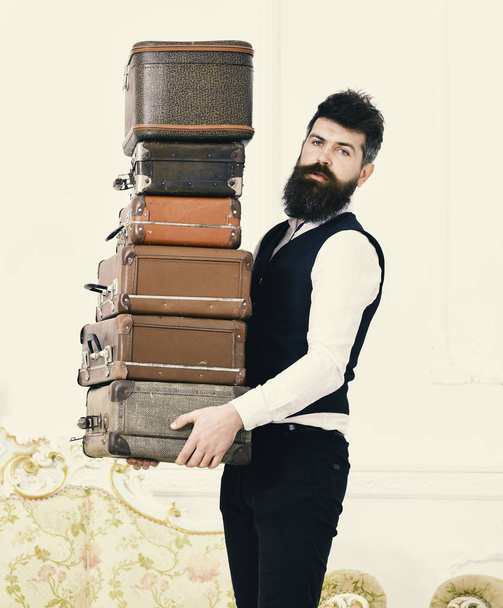 Butler and service concept. Man with beard and mustache wearing classic suit delivers luggage, luxury white interior background. Macho, elegant porter carries heavy pile of vintage suitcases - Photo, Image