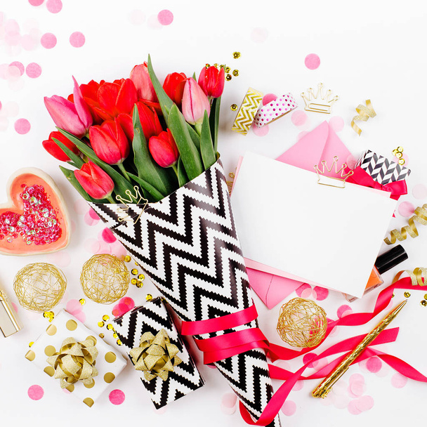 Pink and Gold Styled Desk with Florals. Pink tulips in black and white stylish wrapping paper, gifts, cosmetics and female accessories with confetti on white background - 写真・画像