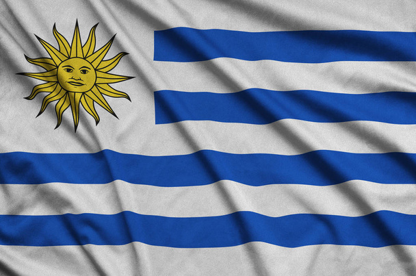 Флаг Уругвая изображен на спортивной ткани со множеством складок. Спортивная команда размахивает баннером
 - Фото, изображение