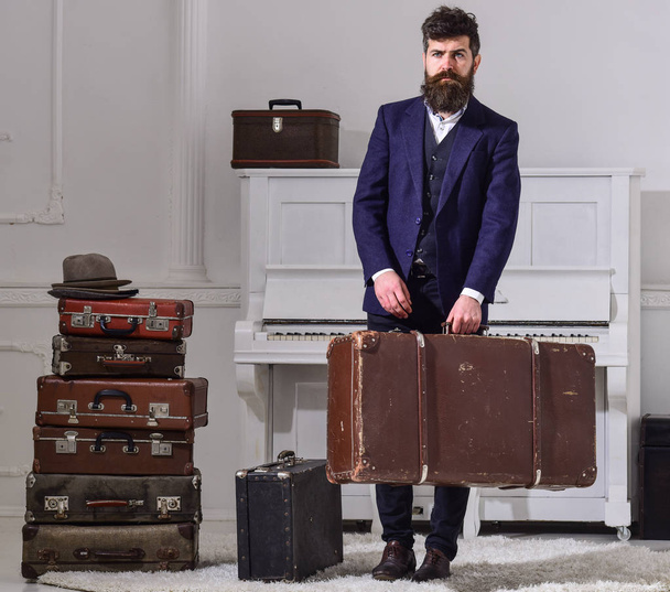Macho elegant on strict face stands near pile of vintage suitcase, holds suitcase. Man, traveller with beard and mustache with luggage, luxury white interior background. Baggage delivery concept - Foto, immagini