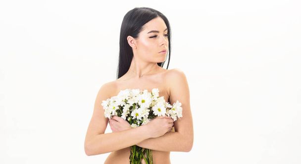 Girl on calm face stands naked and holds chamomile flowers in front of chest. Lady covers breasts with flowers, isolated on white. Woman with smooth healthy skin looks attractive. Skin health concept - Foto, Imagem