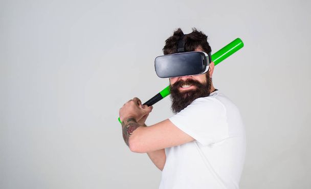 Bearded gamer with agitated look training batting skills, simulation game concept. Man with stylish beard posing with baseball bat isolated on gray background. Bearded man in VR headset doing sports - Foto, afbeelding