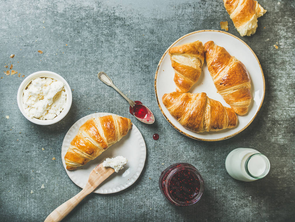 Freshly baked croissants with raspberry jam, cream cheese and milk in bottle over grey concrete background - Foto, Imagen