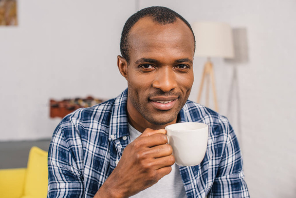 jonge Afro-Amerikaanse man houden koffiekopje en glimlachend op camera - Foto, afbeelding