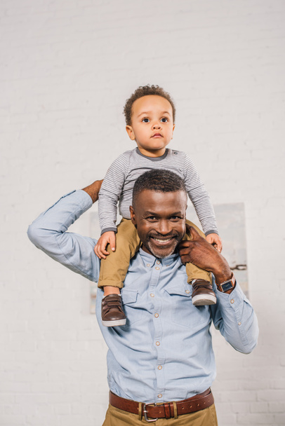 happy african american grandfather carrying little grandchild on neck and smiling at camera - Photo, Image