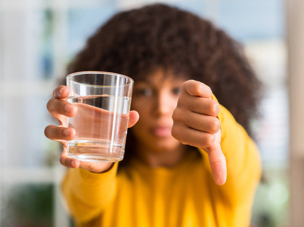 Mujer afroamericana bebiendo un vaso de agua en casa con la cara enojada, signo negativo que muestra aversión con los pulgares hacia abajo, concepto de rechazo
 - Foto, imagen