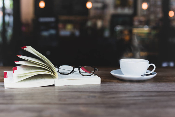 Επιλεγμένο εστίαση της φλιτζάνι καφέ με ανοιχτό βιβλίο και γυαλιά στο ξύλινο τραπέζι σε ματ χρώμα - Φωτογραφία, εικόνα