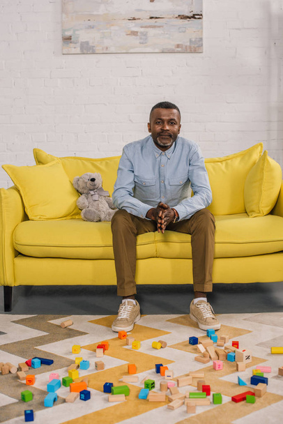 senior african american man looking at camera while sitting on yellow sofa in room with colorful blocks on carpet - Foto, Bild