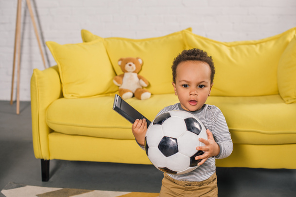 adorable african american toddler holding soccer ball and remote controller at home - Фото, изображение