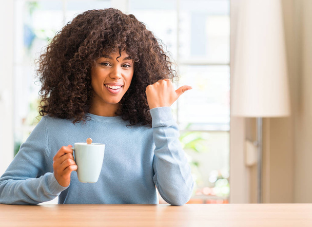 Afro-amerikai nő, egy csésze kávé otthon tartja a kéz és az ujjak felfelé mutató boldog arc mosolyogva - Fotó, kép