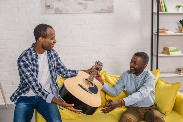 african american father and adult son holding acoustic guitar at home - Fotografie, Obrázek