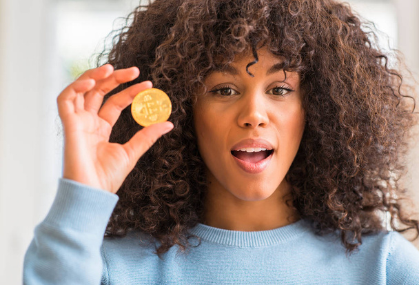 African american woman holding golden bitcoin cryptocurrency at home scared in shock with a surprise face, afraid and excited with fear expression - Foto, Bild